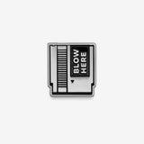 A black and silver soft enamel pin in a retro game cartridge look with the text 'blow here' sitting on a white background.